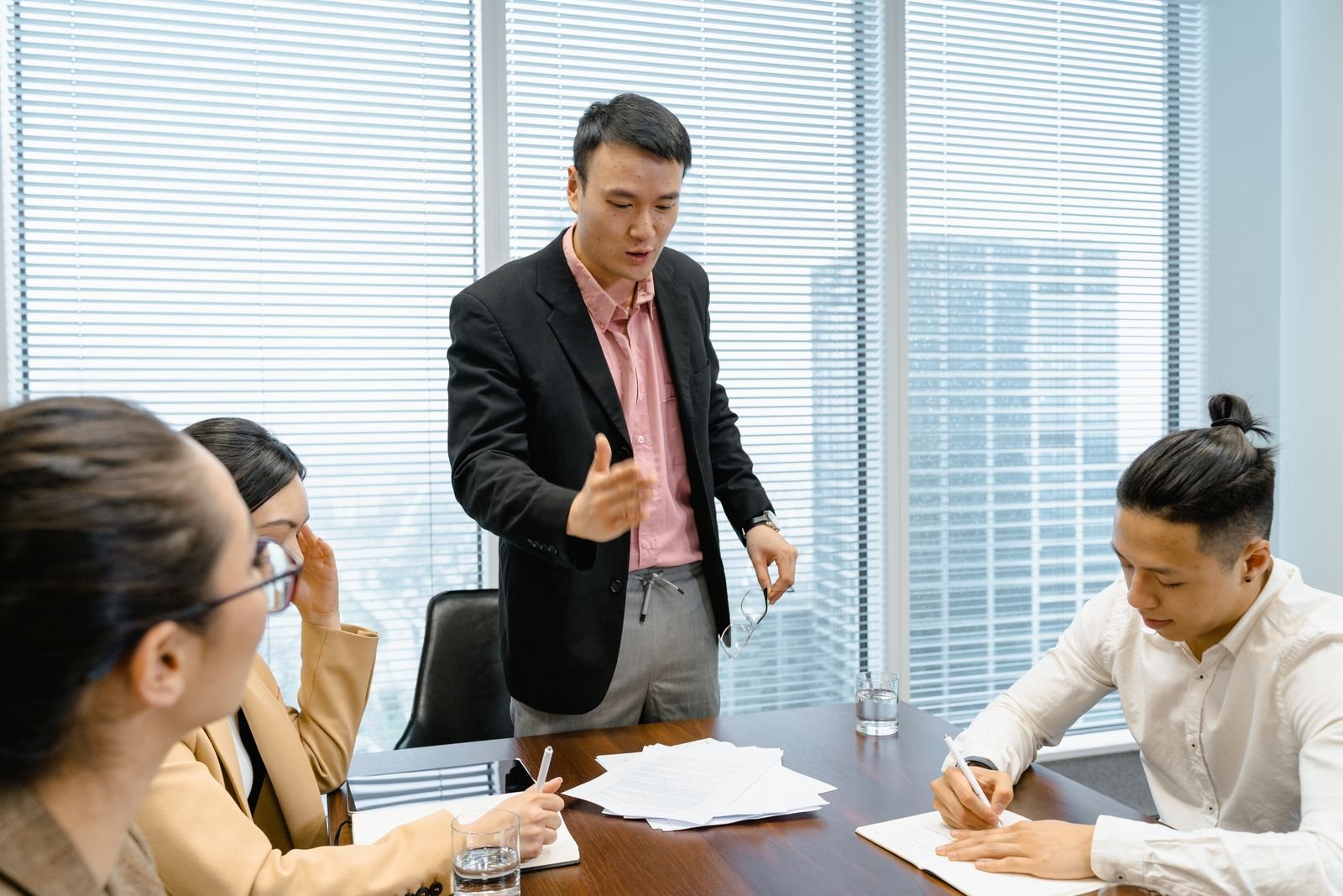 A manager standing over his team in a meeting room, lecturing while his staff look down at their notes